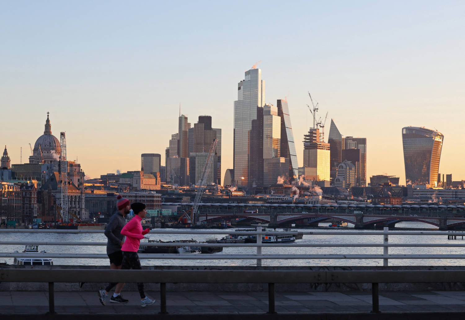 Joggers Run Over Waterloo Bridge, With Skyscrapers Of The City Of London Business District Seen Behind At Sunrise, In London